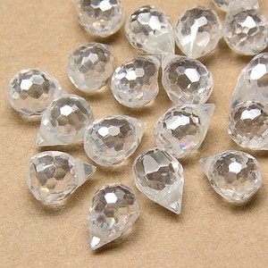 May include: A selection of clear, faceted crystal teardrop beads. The beads have a multifaceted surface that reflects light, creating a sparkling effect. The beads are scattered on a neutral-toned surface.