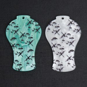 May include: Two decorative pendants with a crane design. One pendant is a marbled teal color, and the other is white. Both pendants have a hole at the top for hanging. The crane design is black and features birds and clouds.