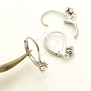 May include: Close-up of three silver-colored leverback earring findings. Each finding has a small, clear, round gemstone at the base. One finding is being held by a pair of tweezers. The findings are on a white background.