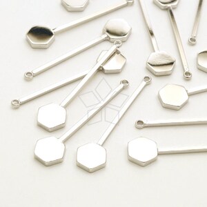 Puede incluir: Un grupo de dijes hexagonales plateados con una varilla plateada unida a cada dije. Los dijes están esparcidos sobre una superficie blanca.