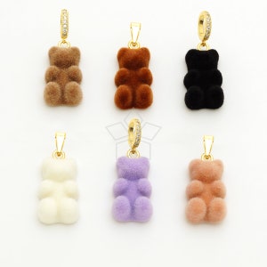 Op de afbeelding: Zes pluche teddybeer bedels in verschillende kleuren, waaronder bruin, zwart, wit, paars en roze. Elke bedel heeft een gouden ring met heldere strass steentjes.
