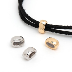 Puede incluir: Primer plano de componentes de joyería. La imagen presenta varias cuentas metálicas de forma ovalada en oro y plata, junto con un cordón negro. Una cuenta dorada está ensartada en el cordón. Las cuentas tienen un acabado liso y pulido.