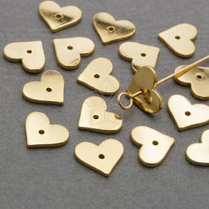 Könnte beinhalten: Eine Sammlung goldfarbener herzförmiger Metallanhänger mit einem Loch in der Mitte. Die Anhänger sind auf einer grauen Oberfläche verstreut.