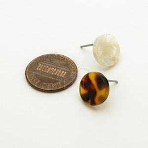 May include: A pair of stud earrings, one with a white and brown tortoiseshell pattern and the other with a white, opaque finish. The earrings are shown next to a US penny for size comparison.
