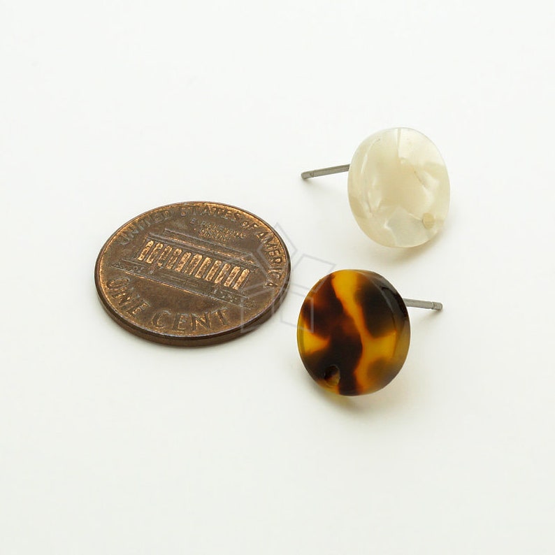May include: A pair of stud earrings, one with a white and brown tortoiseshell pattern and the other with a white, opaque finish. The earrings are shown next to a US penny for size comparison.