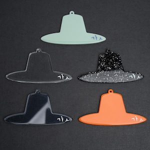 May include: Five hat-shaped ornaments in various colors: light green, clear, black with white speckles, black, and orange. Each ornament has a small hole at the top for hanging and a word in a foreign language.