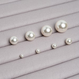 May include: A set of seven white pearl beads of varying sizes, arranged in a row on a grey textured surface.