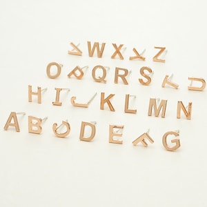 Puede incluir: Un conjunto de pendientes de letras doradas, cada letra del alfabeto está representada. Los pendientes están sobre un fondo blanco.