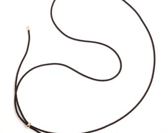 Könnte beinhalten: Dunkelbraune Seilkette mit goldfarbenen Metallakzenten. Die Halskette verfügt über einen Gleitknoten und goldfarbene Kappen an den Enden des Seils, wodurch ein minimalistisches und elegantes Design entsteht. Die Halskette wird vor weißem Hintergrund präsentiert.
