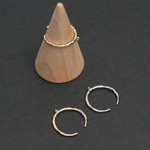 Könnte beinhalten: Zwei gehämmerte Metallringe, einer in Gold und einer in Silber, werden auf einem Holzkegel und einer schwarzen Oberfläche präsentiert. Die Ringe sind offen und haben oben eine kleine Schlaufe.