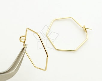 Puede incluir: Un par de pendientes de aro geométricos dorados. Un pendiente está sujeto por unas pinzas plateadas. Los pendientes tienen forma hexagonal, con un diseño sencillo y minimalista. El fondo es blanco liso.
