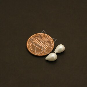 May include: Two pear-shaped, white faux pearls next to a U.S. penny. The pearls have a smooth, lustrous surface. The penny is copper-colored and shows the words "ONE CENT" and "UNITED STATES OF AMERICA".