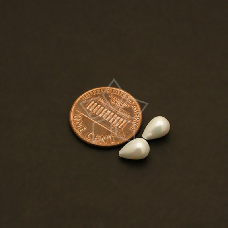 May include: Two pear-shaped, white faux pearls next to a U.S. penny. The pearls have a smooth, lustrous surface. The penny is copper-colored and shows the words "ONE CENT" and "UNITED STATES OF AMERICA".