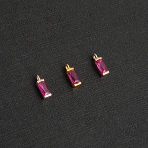 May include: Four gold charms with pink rectangular gemstones. The charms are arranged in a row on a black surface.