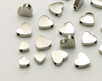 May include: A collection of shiny, silver-colored heart-shaped beads. Each bead has a smooth, reflective surface and a small hole for stringing. The beads vary slightly in size and are scattered against a white background.