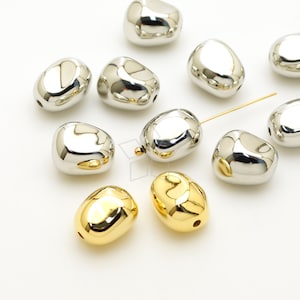 Könnte beinhalten: Eine Sammlung von silbernen und goldenen Metallperlen in Kieselsteinform. Die Perlen sind glatt und glänzend.