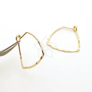 May include: A pair of gold-coloured, geometric earrings. Each earring has a triangular shape with a hammered texture. One earring is held by silver tweezers, while the other is positioned to the right.