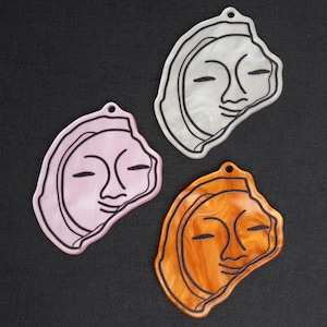 May include: Three face-shaped pendants in pink, white, and orange. Each pendant features a stylized face with closed eyes and a smiling mouth, outlined in black. The pendants have a small hole at the top for hanging.