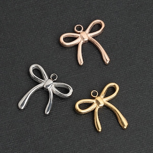 Op de afbeelding: Drie metalen strikcharms in goud, zilver en rosé goud. De charms zijn in de vorm van een strik met een lus aan de bovenkant om aan een ketting of armband te bevestigen.
