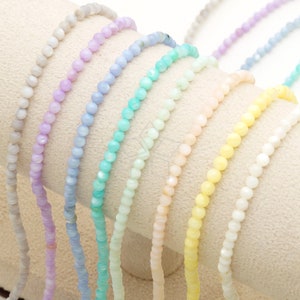 Puede incluir: Primer plano de varios delicados collares de cuentas en una variedad de colores pastel, incluyendo lavanda, azul, verde menta, melocotón, amarillo y blanco. Las cuentas son pequeñas y redondas, ensartadas para crear un aspecto sutil y elegante.
