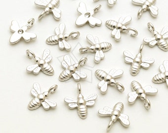 Puede incluir: Una colección de abalorios de abeja plateados con un acabado texturizado. Los abalorios están esparcidos sobre una superficie blanca.