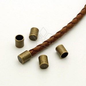 May include: Five antique gold-toned metal cord end caps for leather cord. The cord end caps are cylindrical and have a smooth finish. The cord end caps are shown on a brown leather cord.