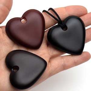 Puede incluir: Tres colgantes en forma de corazón en tonos marrones y negros. Uno es de color burdeos intenso, otro es negro con un cordón negro y el tercero es marrón oscuro. Cada colgante tiene un agujero en la parte superior.