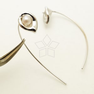 Może przedstawiać: Para srebrnych kolczyków o delikatnym, zakrzywionym wzorze. Każdy kolczyk ma małą, białą perłę osadzoną w kwiatowym oprawie.