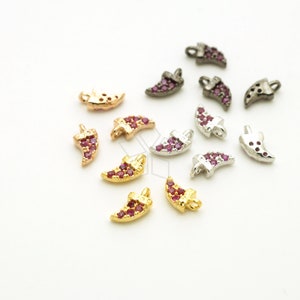 May include: A selection of small, horn-shaped charms in gold, silver, and black metal finishes. Each charm is adorned with small, red gemstones. The charms are scattered on a white surface. The gemstones add a sparkling effect.
