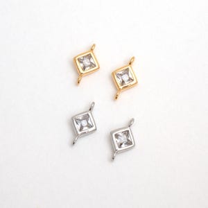 May include: Four small, square-shaped jewelry charms. Two are gold-colored, and two are silver-colored. Each charm features a clear, square-cut gemstone in the center. The charms have small loops at the top for attaching to jewelry.