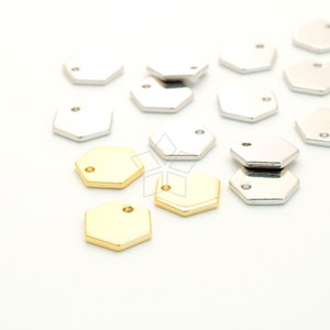 Puede incluir: Una colección de dijes hexagonales plateados y dorados con un pequeño agujero en la parte superior. Los dijes están esparcidos sobre una superficie blanca.