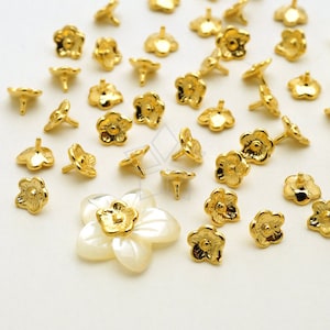 May include: A collection of gold-tone metal flower-shaped embellishments with a small post on the back. The embellishments are scattered on a white surface, with one larger flower-shaped embellishment in the center.