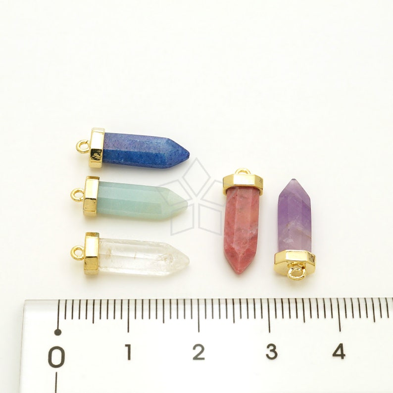 May include: Five gemstone pendants with gold-toned caps and loops. The stones are blue, green, clear, pink, and purple. The pendants are arranged on a white surface with a ruler for scale.