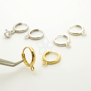 Puede incluir: Un conjunto de pendientes de aro dorados y plateados con un pequeño bucle en la parte inferior. Los pendientes están sobre una superficie blanca.