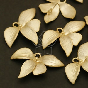 May include: Close-up of several gold-colored metal flower charms. Each charm has three petals and a small loop for attaching to jewelry or other crafts. The charms have a matte finish and are arranged on a dark surface.