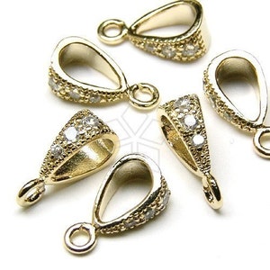 May include: Close-up of several gold-colored pendant bails with a teardrop shape. The bails are encrusted with small, clear gemstones along the edges, and each has a small loop at the top for attaching to a chain or cord. Jewelry findings.