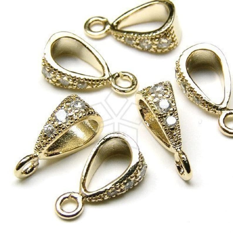 May include: Close-up of several gold-colored pendant bails with a teardrop shape. The bails are encrusted with small, clear gemstones along the edges, and each has a small loop at the top for attaching to a chain or cord. Jewelry findings.
