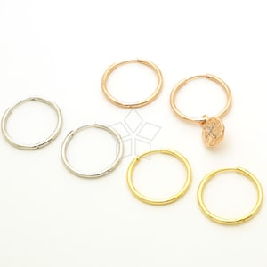 May include: A selection of hoop earrings in silver, rose gold, and gold finishes. One rose gold earring has a small, textured charm. The earrings are presented on a white background, highlighting their simple design and various metallic colours.