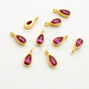May include: Gold-colored teardrop-shaped pendants with a vibrant magenta-colored stone center. The pendants have a textured, beaded border and a small loop at the top for attaching to a chain or other jewelry component. The pendants are scattered on a white surface.