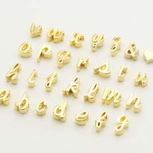 Puede incluir: Un conjunto de abalorios de letras doradas en una fuente cursiva. Las letras incluyen a, b, c, d, e, f, g, h, i, j, k, l, m, n, o, p, q, r, s, t, u, v, w, x, y y z. También hay un ampersand y un abalorio en forma de corazón.