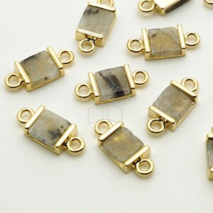 Pode incluir: Conectores de joias retangulares dourados com incrustação de pedra natural. Os conectores têm uma argola em cada extremidade para fixação. A pedra apresenta uma mistura de cores cinza e bege.