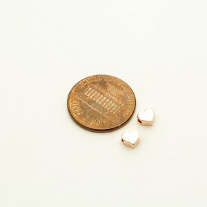 May include: Two small gold-colored heart-shaped charms with a hole in the center. The charms are shown next to a US penny for size comparison.