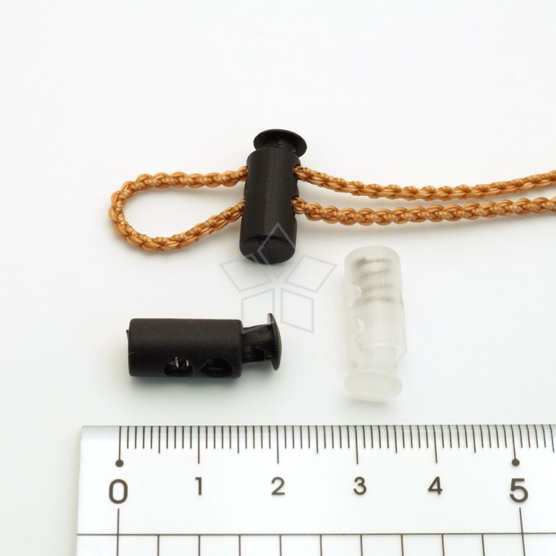 May include: Two black plastic cord locks with a brown cord threaded through one of them. A clear plastic cord lock is also shown. A ruler is in the background.