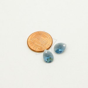 May include: Two blue teardrop-shaped crystal beads with a small hole at the top, next to a penny. The beads have a gradient color, transitioning from a lighter blue at the top to a darker blue at the bottom. The penny is copper-colored.