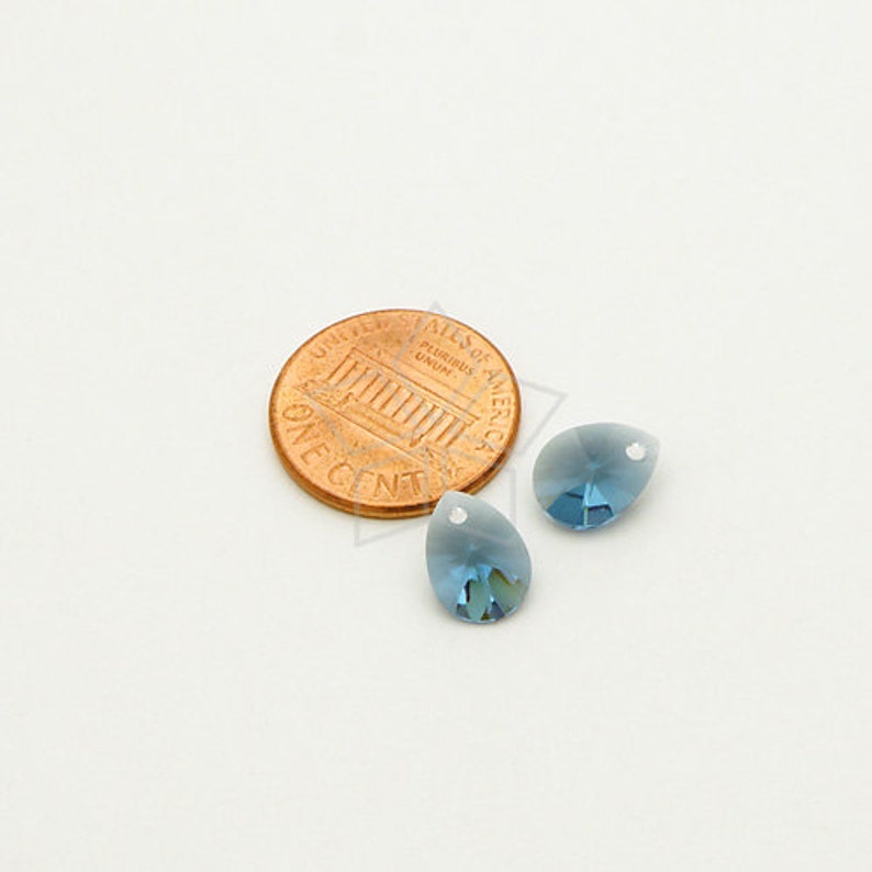May include: Two blue teardrop-shaped crystal beads with a small hole at the top, next to a penny. The beads have a gradient color, transitioning from a lighter blue at the top to a darker blue at the bottom. The penny is copper-colored.
