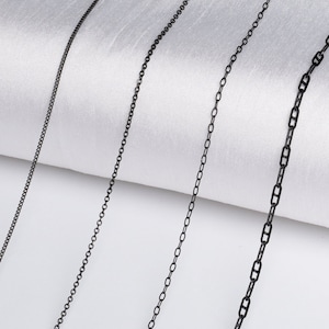 May include: Three black chain necklaces of different widths and link styles, laid out on a white fabric background.