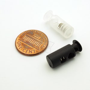 May include: Two plastic cord locks, one black and one clear, next to a US penny. The black cord lock has two holes for threading cord. The clear cord lock has a spring inside.