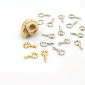 May include: An assortment of silver and gold-colored screw eye pins, scattered on a white surface. A small, off-white skull bead is also present. These small metal eye pins are used for jewelry making and crafts.