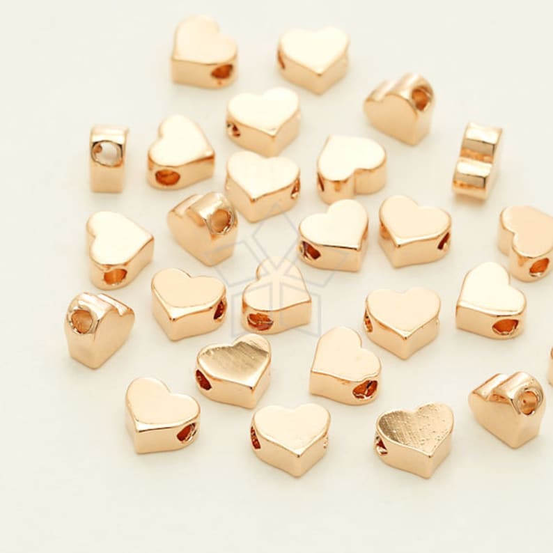 May include: A collection of gold-toned heart-shaped beads with a hole through the center. The beads are scattered on a white surface.