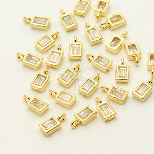 May include: Gold-colored rectangular pendants with a clear, rectangular stone set in the center. Each pendant has a small loop at the top for attaching to a chain or other jewelry component. The pendants are scattered across a white surface.